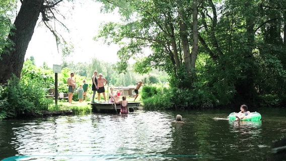 An einem Steg am Flussufer tummeln sich badende Kinder und Erwachsene. © Screenshot 