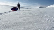 Eine junge Frau in Schneeausrüstung zieht einen Schlitten mit Expeditionsgepäck durch eine weite Schneelandschaft. © Screenshot 