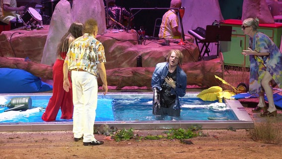 Spielszene aus dem Stück "Blue Skies". Einer der Schauspieler steht in einem Swimmingpool, welcher Teil des Bühnenbilds ist. © Screenshot 