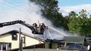 Zwei Feuerwehrleute löschen per Drehleiter den Dachstuhl eines Hauses. © Screenshot 