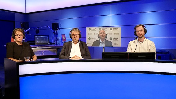 Zwei Männer und eine Frau sitzen in einem Radiostudio. © Screenshot 