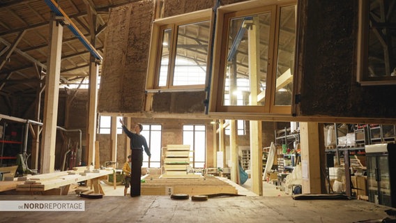 Eine Person steht auf einer Baustelle in einem unfertigen Haus. Durch  die Fensteröffnungen dringt helles Licht. Das Baumaterial ist hell, wie frisches Holz. © Screenshot 