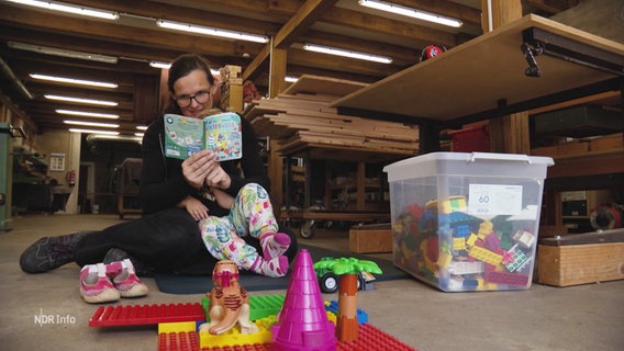 Tischlerin Johanna Röh sitzt in ihrer Tischlerei auf dem Boden und liest ihrem Kind aus einem Buch vor. Um sie herum stehen verschiedene Spielsachen. © Screenshot 