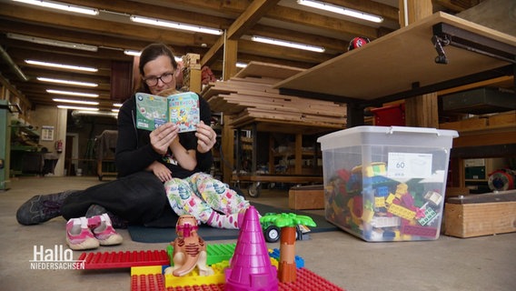 Tischlerin Johanna Röh sitzt in ihrer Tischlerei auf dem Boden und liest ihrem Kind aus einem Buch vor. Um sie herum stehen verschiedene Spielsachen. © Screenshot 