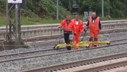 Gleisarbeiter der Deutschen Bahn in oranger Arbeitskleidung und mit Arbeitsgerät auf den Schienen. © Screenshot 