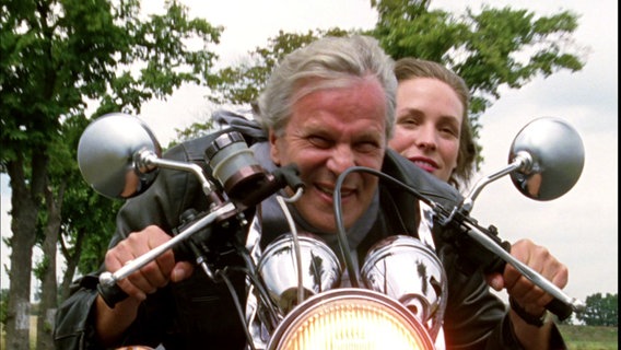 Ein Mann mit grauen Haaren, die im Wind wehen fährt Motorrad. Hinter ihm sitzt eine junge Frau auf dem Motorrad, auch ohne Helm. © Screenshot 