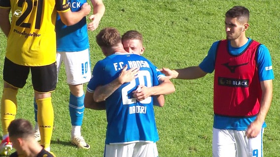 Zwei Spieler des FC Hansa Rostock umarmen sich auf einem Spielfeld. © Screenshot 