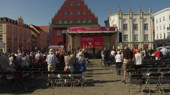 Auf einem marktplatz in Greifswald ist eine Bühne aufgebaut, davor stehen Senschen und beten. © Screenshot 