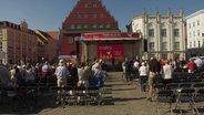 Auf einem marktplatz in Greifswald ist eine Bühne aufgebaut, davor stehen Senschen und beten. © Screenshot 