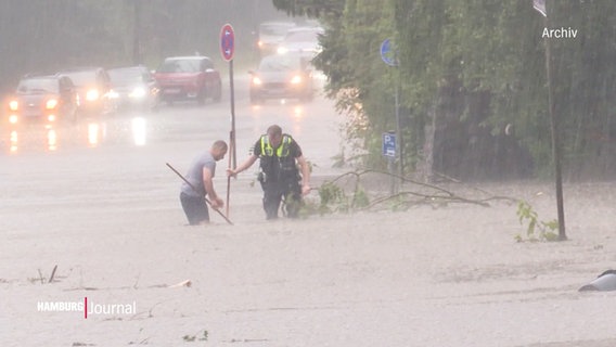 Ein Polizist und ein weiterer Mann versuchen eine überflutete Straße von Debris freizumachen. © Screenshot 