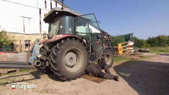 Ein Landmaschinenschlosser liegt unter einem Schlepper und repariert dessen Unterboden. © Screenshot 