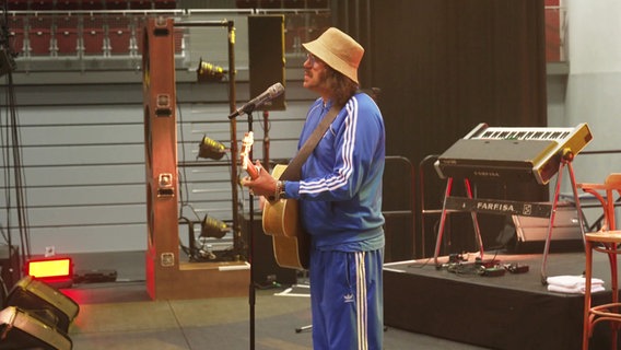 Ein Mann mit blauem Trainingsanzug und Hut steht mit einer Gitarre am Mikrofon © Screenshot 