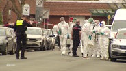 Menschen in weißen Schutzanzügen stehen hinter polizeilichem Absperrband auf einer Straße. © Screenshot 