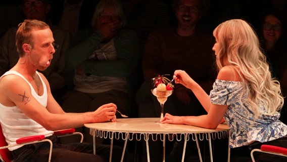 Szene aus dem Theaterstück "Aus unseren Feuern": Eine Frau und ein Mann sitzen an einem Tisch und teilen sich einen Eisbecher. © Screenshot 