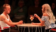 Szene aus dem Theaterstück "Aus unseren Feuern": Eine Frau und ein Mann sitzen an einem Tisch und teilen sich einen Eisbecher. © Screenshot 