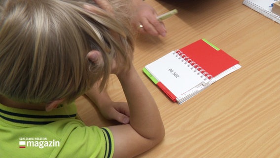 Ein Junge sitzt an einem Tisch und stützt seinen Kopf mit einer Hand. Vor ihm liegt ein Ringbuch, das eine kurze Zahlenfolge zeigt. © Screenshot 