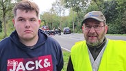 Ein junger und ein älterer Mann stehen auf einer Landstraße. Im Hintergrund ist ein Autounfall zu sehen. © Screenshot 