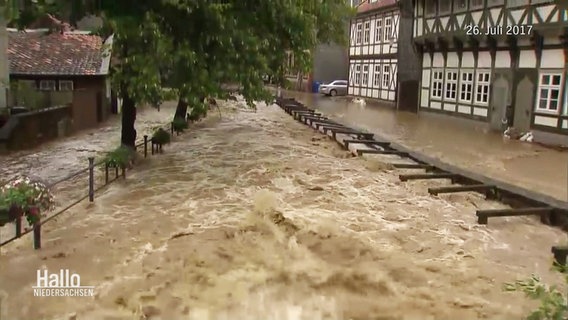 Ein Fluss ist über die Ufer getreten. Das Wasser steht auf Gehweg und Straße und droht in Häuser zu laufen. © Screenshot 