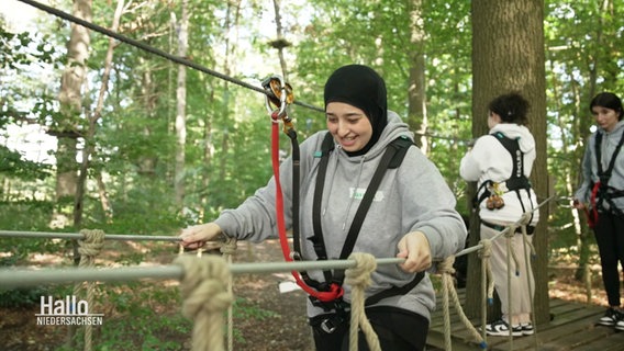 Ein Mädchen mit grauem Sweatshirt und Kopftuch klettert lächelnd über eine Seilbrücke © Screenshot 