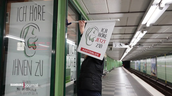 Eine Frau hält eine Fahne in einer U-Bahn-Station. Das Banner enthält die Aufschrift „Ich höre jetzt zu! Kostenlos“. © Screenshot 