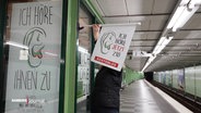 Eine Frau hält eine Fahne in einer U-Bahn-Station. Das Banner enthält die Aufschrift „Ich höre jetzt zu! Kostenlos“. © Screenshot 