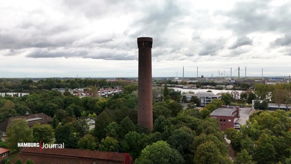 Ein hoher Schornstein, umgeben von Bäumen und einem weiten Blick auf die Landschaft. Im Hintergrund sind Windräder und Industrieanlagen sichtbar. © Screenshot 