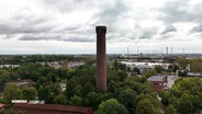 Ein hoher Schornstein, umgeben von Bäumen und einem weiten Blick auf die Landschaft. Im Hintergrund sind Windräder und Industrieanlagen sichtbar. © Screenshot 