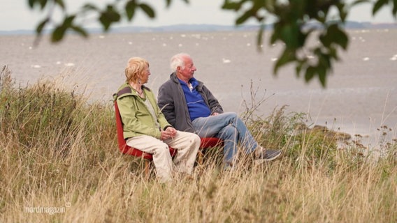 Ein älteres Paar sitzt auf zwei roten Stühlen am Wasser. © Screenshot 