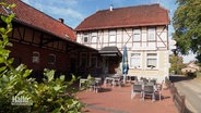 Eine Gaststätte in einem Fachwerkhaus im Sonnenschein. Vor dem Haus eine Terrasse mit Tischen und Stühlen. © Screenshot 