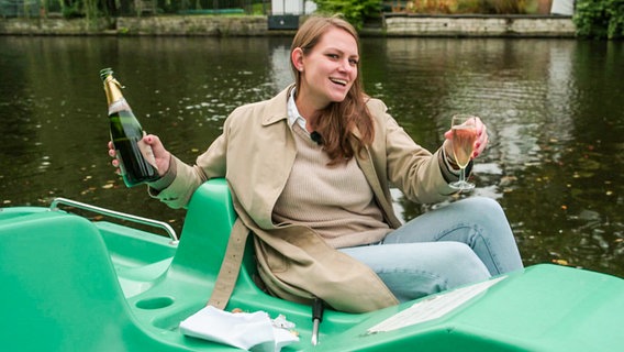 Reporterin Céline in einem Tretboot auf der Alster mit einer Champagnerflasche und Glas. (extra 3 vom 10.10.2024 im Ersten) © NDR 