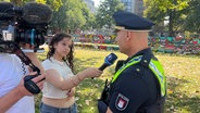 Eine junge Frau interviewt einen Polizisten im Freien. Ein Kameramann filmt das Gespräch. Im Hintergrund sind Bäume und eine bunt bemalte Wand zu sehen. © Screenshot 