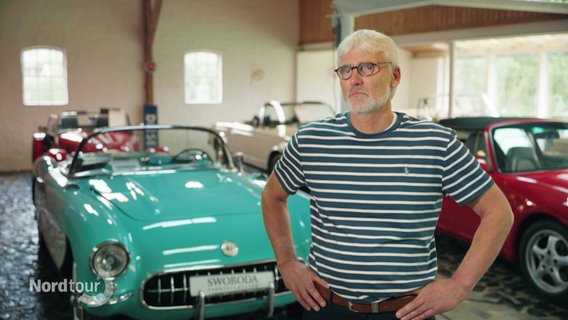 Jürgen Swoboda steht in seiner Halle vor verschiedenen Oldtimer Wagen. © Screenshot 