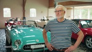 Jürgen Swoboda steht in seiner Halle vor verschiedenen Oldtimer Wagen. © Screenshot 