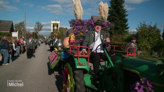 Ein Mann in Tracht sitzt auf einem geschmückten Traktor. © Screenshot 