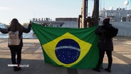 Zwei Menschen stehen mit einer Brasilien-Flagge am Hamburger Hafen, im Hintergrund passiert ein Marine-Schulschiff die Elbe. © Screenshot 