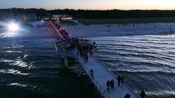 Zahlreiche Menschen laufen bei einbrechender Dunkelheit über die neu eröffnete und hell illuminierte Seebrücke in Prerow auf die Ostsee hinaus. © Screenshot 