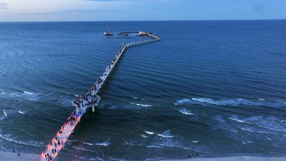 Zahlreiche Menschen laufen bei einbrechender Dunkelheit über die neu eröffnete und hell illuminierte Seebrücke in Prerow auf die Ostsee hinaus. © Screenshot 