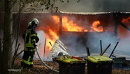 Ein Feuer wütet an einem Haus und zwei Rettungskräfte versuchen den Brand zu löschen. © Screenshot 