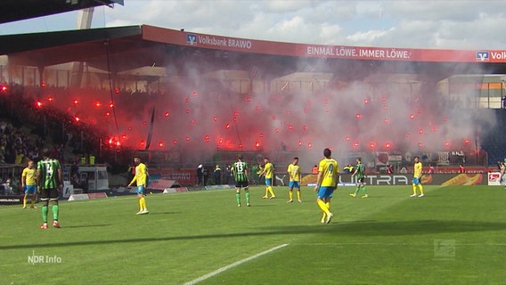 Szene bei einem Fußballspiel: Randalierer zünden Pyrotechnik auf den Tribünen, im Vordergrund Fußballer auf dem Rasen. © Screenshot 