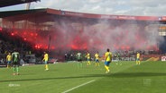 Szene bei einem Fußballspiel: Randalierer zünden Pyrotechnik auf den Tribünen, im Vordergrund Fußballer auf dem Rasen. © Screenshot 