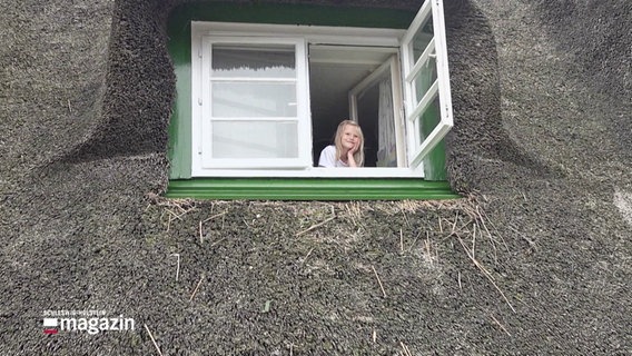 Ein blondes Mädchen im Grundschulalter schaut aus dem offenen Fenster eines reetgedeckten Hauses. © Screenshot 