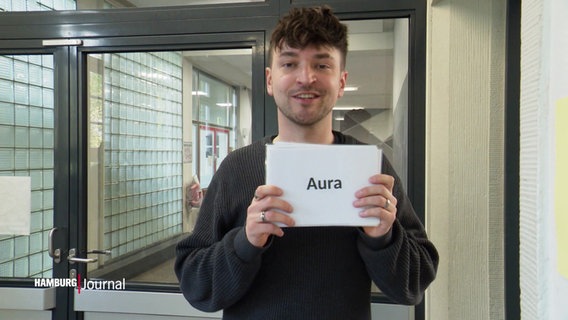 NDR Reporter Tristan Dück hält ein Schildt mit dem Wort "Aura" in der Hand. © Screenshot 