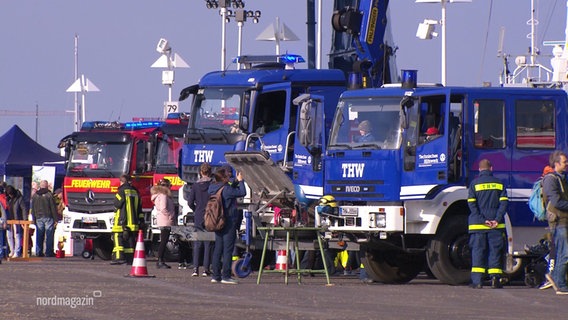 Ein Feuerwehrwagen sowie zwei blaue Wagen des THW stehen in einer Reihe und werden von Besucher*innen fotografiert. © Screenshot 