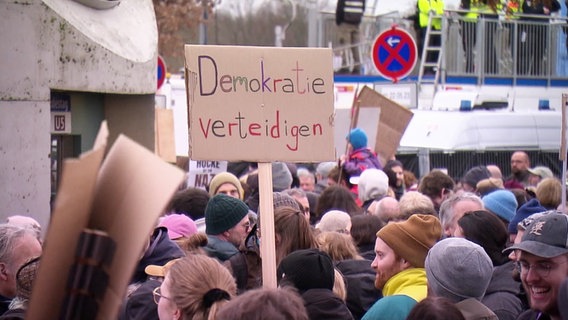 Auf einer Demonstration wird ein Schild hochgehaltenauf dem steht "Demokratie verteidigen!". © Screenshot 