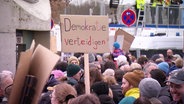 Auf einer Demonstration wird ein Schild hochgehaltenauf dem steht "Demokratie verteidigen!". © Screenshot 