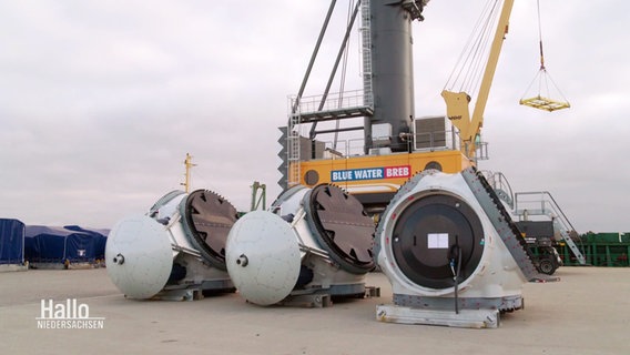 Mehrere Köpfe von Windkrafträdern stehen ohne Rotorblätter auf einer Lagerfläche in einem kleinen Hafen vor einem Kran. © Screenshot 