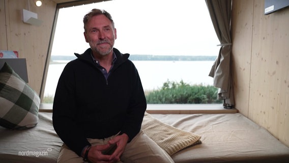 Markus Wolff sitzt vor einem großen Fenster mit Aussicht auf einen See in einem zum Campen umgebauten Tiny House. © Screenshot 