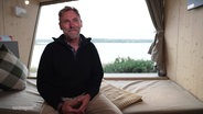 Markus Wolff sitzt vor einem großen Fenster mit Aussicht auf einen See in einem zum Campen umgebauten Tiny House. © Screenshot 