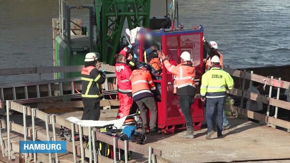 Mithilfe mehrerer Rettungskräfte wird ein schwerverletzter Hafenarbeiter auf einer Trage mit einer Ein-Personen-Kran-Gondel geborgen. © Screenshot 