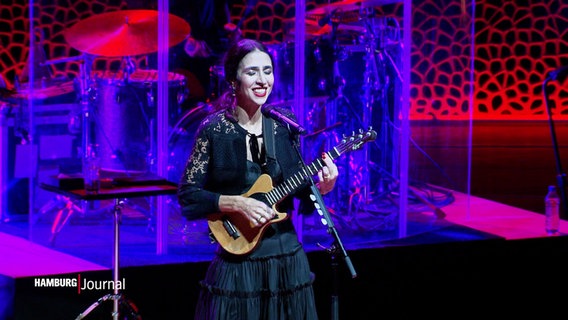 Die brasilianische Sängerin Marisa Monte spielt auf einer kleinen Gitarre auf der Bühne der Elbphilharmonie und lächelt. © Screenshot 